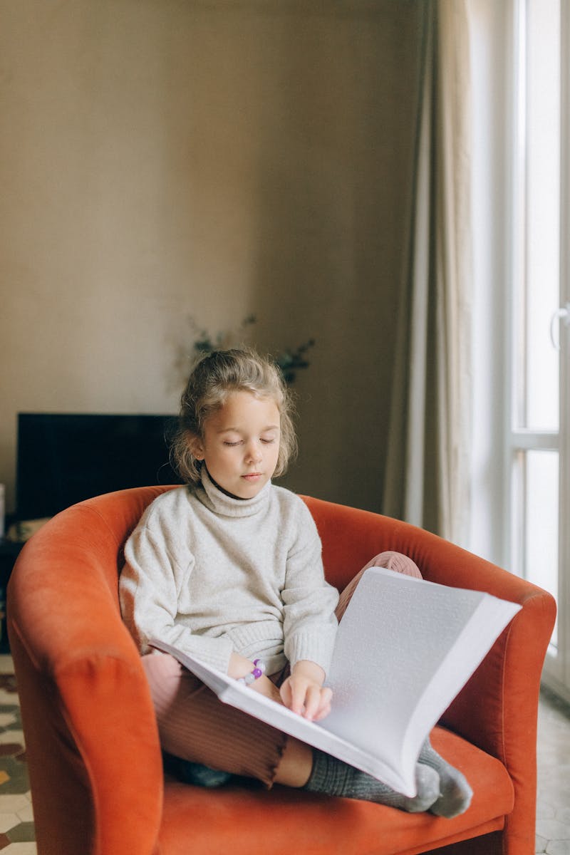 A young girl with closed eyes reads a Braille book at home, highlighting learning and disability.